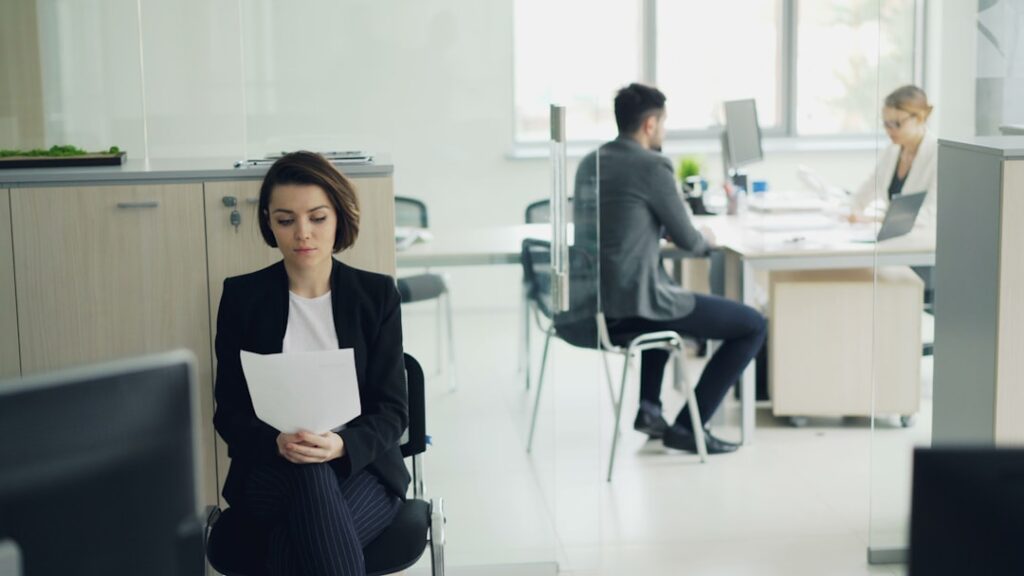 A candidate in professional clothing sits across a desk from an interviewer in a bright, modern office.