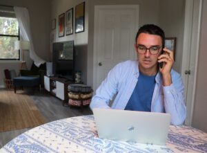 A woman works at a desk with a laptop and notepad in a sunlit living room, with a child playing in the background.